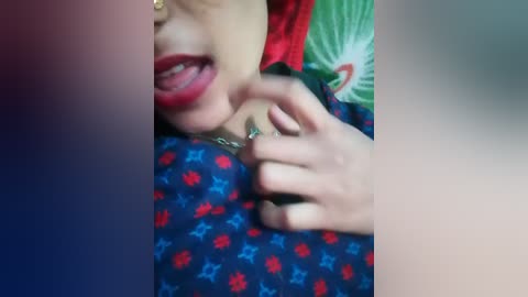 Video of a young woman with light skin, wearing red lipstick, a black and red star-patterned dress, and a green scarf. She playfully sticks her tongue out and points with her right hand.