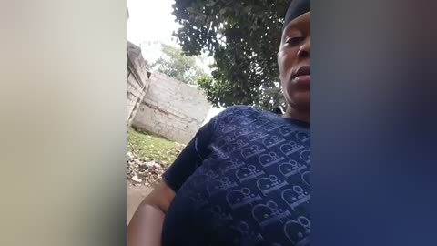 Video of a person wearing a dark blue t-shirt with white icons, standing in a sunlit outdoor area with greenery, a concrete wall, and scattered rocks in the background.