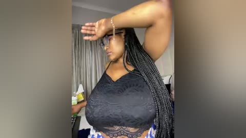Video of a dark-skinned woman with braided hair in a black lace top, adjusting glasses in a dimly lit bedroom with a bed and curtains.
