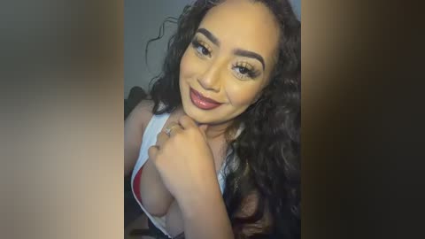 Video of a smiling woman with long, curly black hair, wearing a white tank top revealing cleavage, in front of a blurred background.