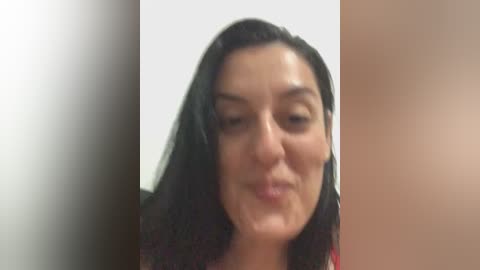 Video of a middle-aged woman with medium-length black hair, light skin, and a neutral expression. She is wearing a red top. The background is a plain white wall.