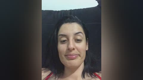 A video of a smiling, fair-skinned woman with dark hair and minimal makeup, wearing a red top, reclining on a black couch with a blurred background.