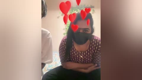 A video of a South Asian woman in a bathroom, wearing a black mask, a purple patterned shirt, and black pants, with red heart emojis obscuring her face.