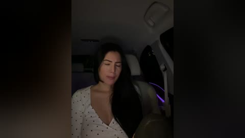 Video of a woman with long black hair, wearing a white polka dot dress, leaning back in a dimly lit car seat, eyes closed, smiling.