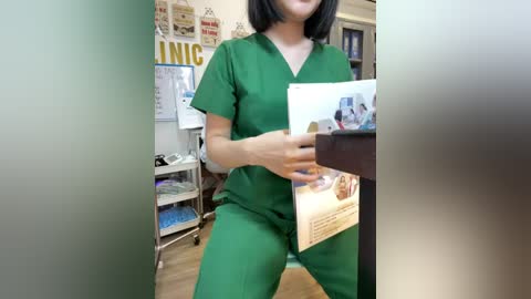 Video of an Asian woman with straight black hair, wearing green scrubs, holding a magazine, standing in a medical office with shelves and posters in the background.