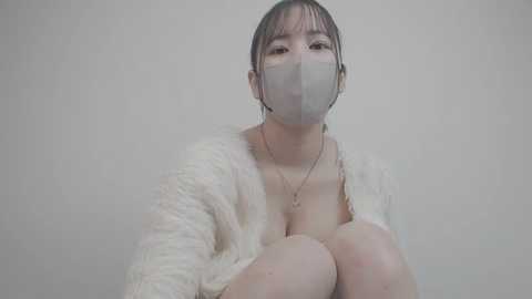 Media: Video of an Asian woman with light skin, wearing a white fluffy cardigan, gray surgical mask, and a delicate gold necklace. She sits against a plain, light gray background, her expression neutral.