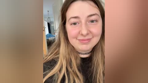 Video of a young woman with fair skin and long, wavy, blonde hair, wearing a black choker necklace, smiling, standing in a dimly lit room with a blue couch and a chandelier visible in the background.