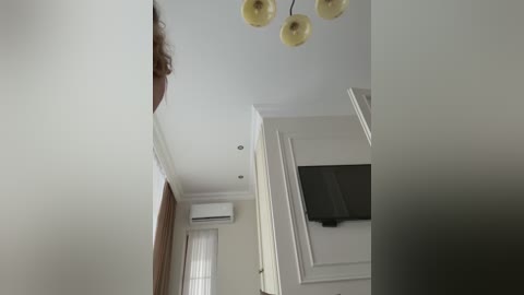 Video of a modern, minimalist room with white walls, a large flat-screen TV mounted on a white panel, a chandelier with yellow shades, and a white air conditioner.