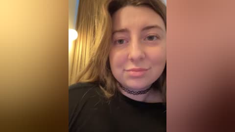 Video of a young woman with light skin, straight brown hair, and a black choker, standing in a beige room with a warm light source.