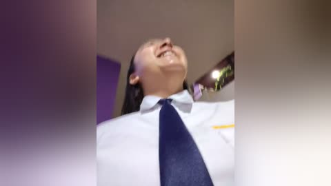 Video of a smiling person in a white dress shirt and navy tie, with a blurred background featuring a wooden ceiling and purple lighting.