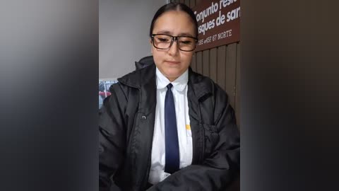 Video of a middle-aged Latina woman with medium skin tone, dark hair in a ponytail, wearing black-framed glasses, a white shirt, dark tie, and a black jacket, standing in a dimly lit hallway with Spanish signage.
