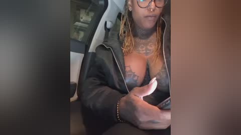 A video of a black woman with braids, tattoos, and glasses, wearing a black leather jacket, partially unzipped, revealing cleavage, sitting in a car.
