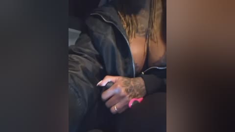 A video showing a woman in a black leather jacket, with a tattooed hand touching her breast, revealing a cleavage. The background is blurred, focusing on the intimate gesture.