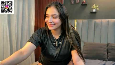 Video of a young woman with long dark hair, smiling, wearing a black t-shirt, seated on a bed in a modern bedroom with grey headboard and dark walls.