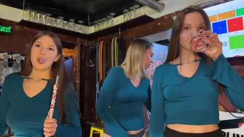 Video of three young women with straight brown hair, wearing teal tops, playfully drinking from straws inside a cozy bar with wooden walls, shelves, and colorful signage.