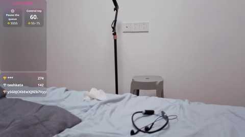 Video of a minimalist bedroom with a white wall, a small gray stool, and a bed with light blue sheets. A black microphone cable lies on the bed.