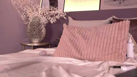 A video of a cozy bedroom with a white bed, a pink knitted throw pillow, a textured silver vase with white flowers, and a bottle of lotion on a bedside table.