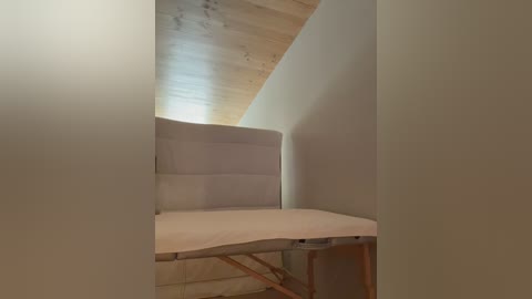 Media: A video of a small, simple room with a slanted wooden ceiling, beige walls, and a white massage table with wooden legs, set against a dimly lit backdrop.