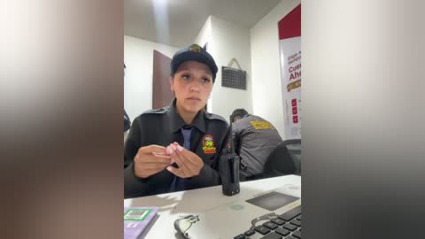 Video of a female police officer in uniform, holding a pink cell phone, seated at a desk in a dimly lit office.