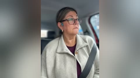 Video of a middle-aged woman with glasses and gray hair, wearing a beige coat, gray seatbelt, and maroon top, seated in a car with blurred background.