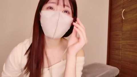 A video of a young Asian woman with long dark hair, wearing a white mask, white top, and talking on a phone indoors.