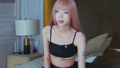 Video of a young, slender Asian woman with long pink hair, wearing a black crop top and high-waisted pants, sitting on a bed in a dimly lit room with a white lamp and yellow curtains.