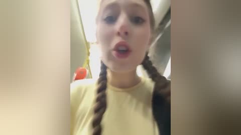 A video of a young Caucasian woman with light skin, wearing a yellow shirt, with long braided hair. She's in an indoor setting, possibly a bathroom, with blurred background objects.