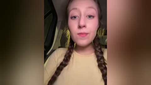 A video of a young girl with light skin, brown hair in braids, wearing a beige top, sitting in a car with a brown interior.