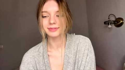 Video of a young, fair-skinned woman with light brown hair, wearing a gray robe, smiling with a lip ring, in a simple room with a wall-mounted light fixture.