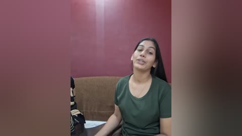 Video of a woman with long dark hair, wearing a green shirt, sitting at a table with a brown cushion, in a room with red walls, lens flare visible.