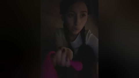 A dimly lit video shows a young woman with dark hair, wearing a white shirt and black apron, holding a pink object in her hand, against a shadowy background.