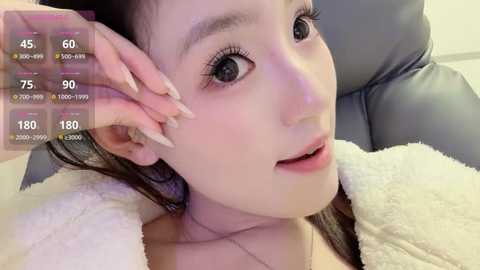 Video of an Asian woman with light skin, long straight hair, and large eyes, wearing a fluffy white robe, lying on a black leather chair, with virtual health and fitness metrics displayed.