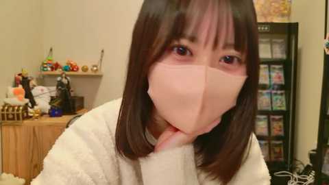 A video of an Asian woman with straight, shoulder-length brown hair, wearing a white fuzzy sweater and a pink face mask, in a cluttered room with a wooden shelf, a refrigerator, and colorful kitchen items in the background.