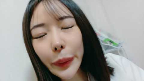 Video of an Asian woman with long, straight black hair, light skin, and full lips. She has her eyes closed and a slight smile. Background shows a blurred white wall and a white bed.