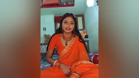 Video of a South Asian woman in an orange sari with intricate embroidery, wearing red lipstick and jewelry, seated on a bed in a modest room with a wooden door and simple furnishings.