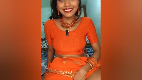 Video of a South Asian woman with medium brown skin, wearing a bright orange saree and matching top, adorned with jewelry, sitting on a bed with a patterned blanket, smiling.