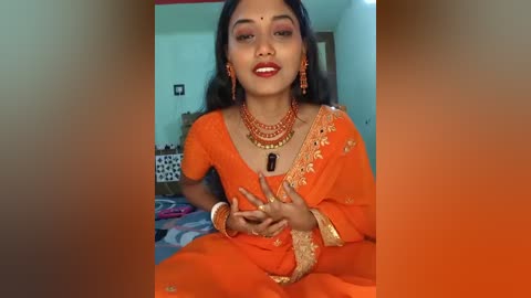 Video of a South Asian woman with medium skin tone and dark hair, dressed in an orange sari adorned with gold embroidery, sitting indoors. She wears red jewelry and makes a peace sign with her hands.