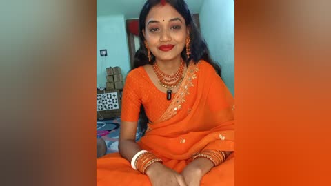 Video of a South Asian woman in a bright orange sari, adorned with gold jewelry, seated in a modest home with white walls and a patterned rug.