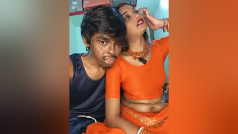 Video of a young Indian couple; man in black sleeveless shirt, woman in orange lace crop top and matching skirt, both smiling.