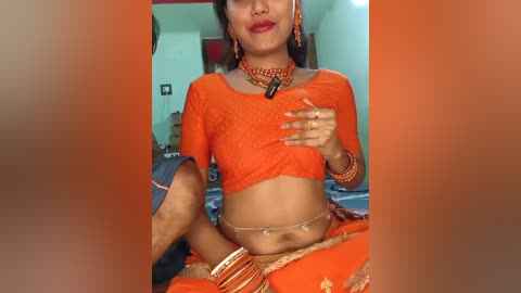 Media: Video of a South Asian woman in an orange saree, adorned with traditional jewelry, sitting on a bed with a blue blanket, wearing a long-sleeved blouse and a gold necklace.