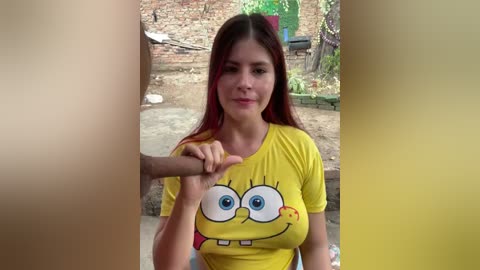 Video of a young woman with long, straight red hair, wearing a yellow SpongeBob shirt, sitting outdoors. She has a neutral expression and is making a thumbs-up gesture. The background features a rustic, sunlit yard with a stone wall and greenery.