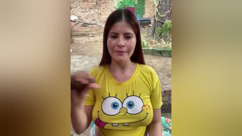 Video of a young woman with long red hair, fair skin, and blue eyes, wearing a yellow SpongeBob shirt. She gestures with her hand. Background shows a brick wall and greenery.