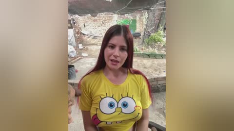 A video of a young woman with long, straight auburn hair, wearing a yellow SpongeBob SquarePants t-shirt. She stands in a rustic, outdoor setting with a stone wall and greenery in the background.