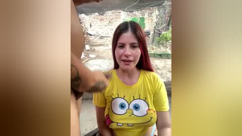 Video of a woman with long red hair, wearing a yellow SpongeBob shirt, sitting in a rustic outdoor setting. A man's erect penis is partially visible in the foreground.