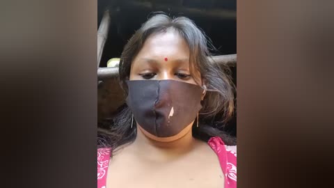 Video of a woman with medium brown skin, long black hair, and a red dot on her forehead, wearing a black surgical mask and a red patterned top. Background is blurred and dark.