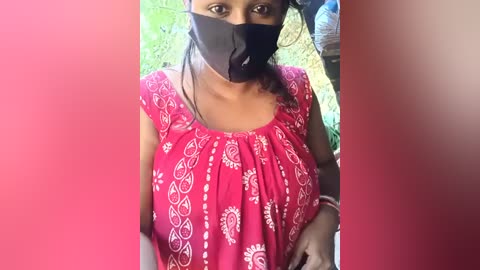 Video of a woman wearing a black face mask, black top, and red patterned shirt, standing outdoors with blurred green foliage and a person partially visible in the background.