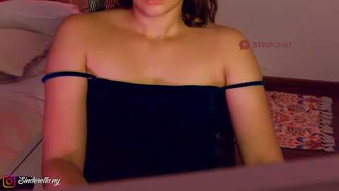 Media: A video of a woman with light skin, dark hair, and a black strapless top, lying on a bed with floral-patterned pillows in a dimly lit room.