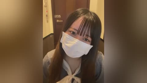 A video of a young Asian woman with long, straight brown hair, wearing a white surgical mask, a grey cardigan, and a white blouse, standing in a hallway with wooden doors labeled \"1205\" and \"1206.\