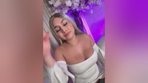 Video of a smiling, light-skinned woman with long, straight blonde hair, wearing a white off-shoulder top, in a room with purple lighting and cherry blossom wallpaper.
