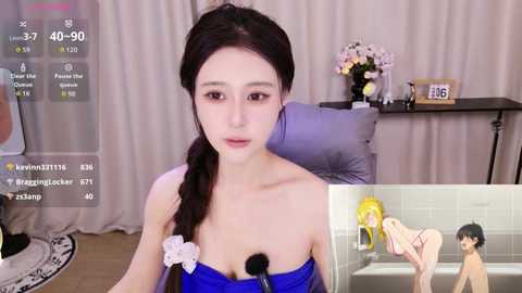 A video of a young East Asian woman with long brown hair in a braid, wearing a strapless blue dress, sitting on a grey chair in a modern room with a flower vase and clock.
